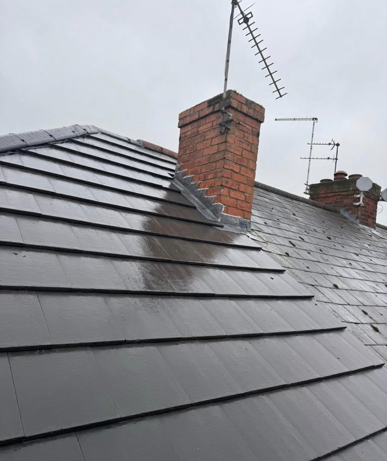 Angled view of a residential roof with gray slate tiles, red brick chimneys, and TV antennas against a cloudy sky