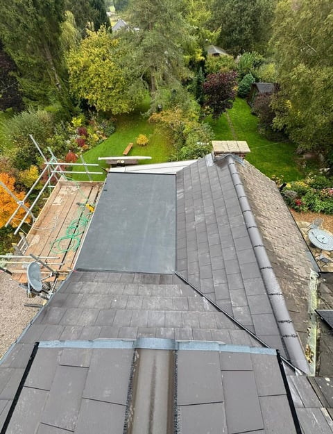 Aerial view of a residential roof with dark shingles under renovation surrounded by lush green garden and trees