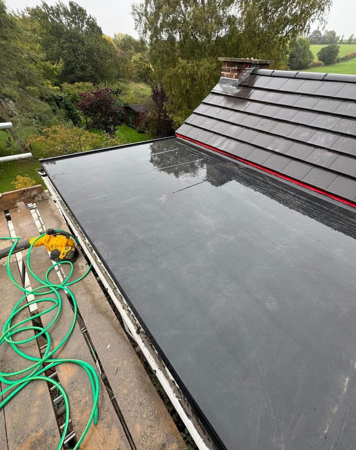 Flat roof with dark membrane and red trim, adjacent to slate-tiled pitched roof, with green garden hose and lush trees in background