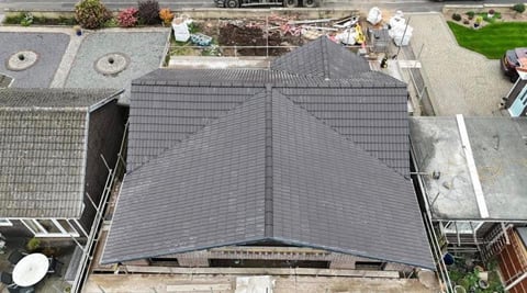 Aerial view of a modern house with a dark gray peaked roof over the center section, surrounded by paved areas and neighboring properties